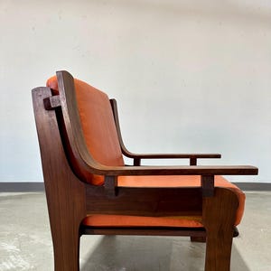May include: A mid-century modern armchair with a dark wood frame and orange leather upholstery. The chair has a unique design with a high back and wide arms.