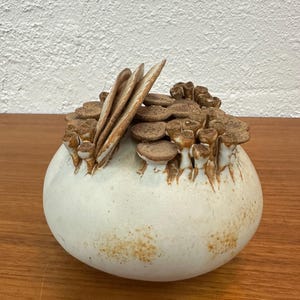 May include: A ceramic, off-white, bulbous vase with a textured surface. The top of the vase features a cluster of brown, mushroom-like shapes and several vertical, leaf-like elements. The vase sits on a wooden surface.