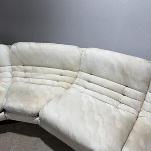 May include: Off-white sectional sofa with a patterned design. The sofa has multiple sections and features a curved shape. The fabric appears to have a subtle wave-like pattern. The cushions have a quilted appearance.