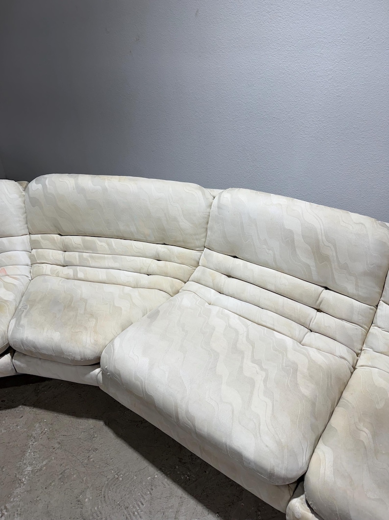May include: Off-white sectional sofa with a patterned design. The sofa has multiple sections and features a curved shape. The fabric appears to have a subtle wave-like pattern. The cushions have a quilted appearance.