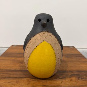 May include: A ceramic penguin figurine with a black head and a yellow body. The penguin's belly is yellow, and the body is decorated with a tan geometric pattern. The figurine is sitting on a wooden surface.