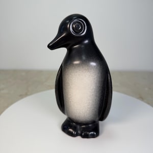 May include: A standing penguin figurine featuring a smooth, matte finish with a gradient color scheme. The exterior is primarily black, transitioning into a soft, light gray or white patch on the chest and belly area. The eyes are circular.