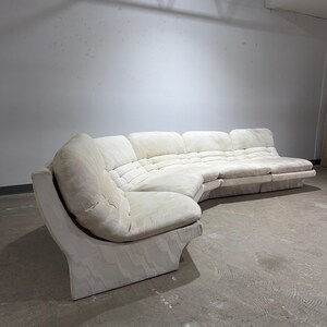May include: A large, off-white sectional sofa with a curved design. The sofa features a tufted seat and back cushions. The base has a wavy pattern. The sofa is in a large, empty room with a concrete floor.