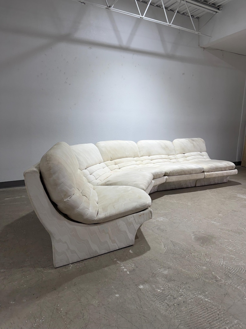 May include: A large, off-white sectional sofa with a curved design. The sofa features a tufted seat and back cushions. The base has a wavy pattern. The sofa is in a large, empty room with a concrete floor.