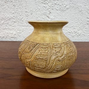 May include: A beige ceramic vase with a textured, carved design. The vase has a rounded body and a flared neck. The carved pattern features a series of horizontal and vertical lines, creating a unique visual texture. The vase sits on a small, circular base.