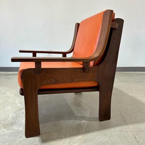 May include: A mid-century modern armchair with a walnut wood frame and orange leather upholstery. The chair has a unique design with a wide, flat armrest and a low back.