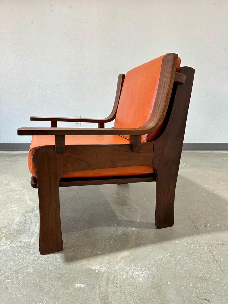 May include: A mid-century modern armchair with a walnut wood frame and orange leather upholstery. The chair has a unique design with a wide, flat armrest and a low back.