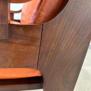 May include: Close-up of a wooden chair with a curved back and orange leather upholstery. The chair is made of dark wood with a smooth finish.