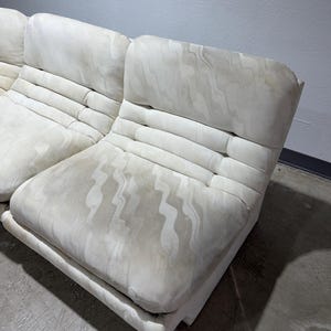 May include: Off-white modular sofa with a patterned fabric. The sofa has a low profile with a rectangular base and a tufted backrest. The fabric has a subtle, wavy design. The sofa is in a room with a concrete floor and a light gray wall.