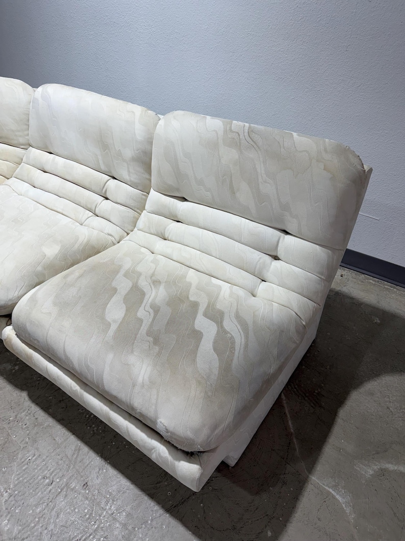 May include: Off-white modular sofa with a patterned fabric. The sofa has a low profile with a rectangular base and a tufted backrest. The fabric has a subtle, wavy design. The sofa is in a room with a concrete floor and a light gray wall.