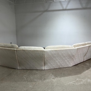 May include: A large, curved sofa with a light-colored, patterned fabric. The sofa has a low profile and a series of connected sections, creating a rounded shape. The fabric has a subtle, wave-like design. The sofa is in a room with a concrete floor.