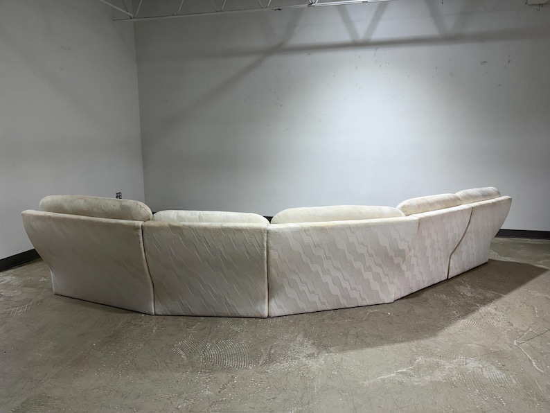 May include: A large, curved sofa with a light-colored, patterned fabric. The sofa has a low profile and a series of connected sections, creating a rounded shape. The fabric has a subtle, wave-like design. The sofa is in a room with a concrete floor.
