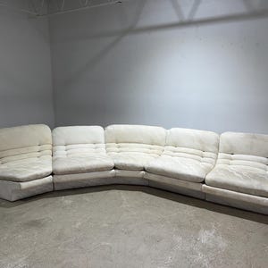 May include: A large, off-white sectional sofa with a curved design. The sofa is composed of multiple modular pieces, each with a tufted back and seat. The fabric appears to be a soft, textured material.