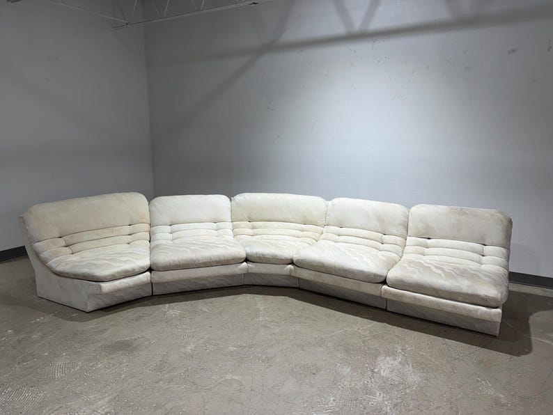 May include: A large, off-white sectional sofa with a curved design. The sofa is composed of multiple modular pieces, each with a tufted back and seat. The fabric appears to be a soft, textured material.