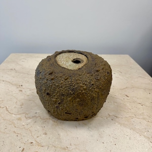 May include: A round ceramic vessel with a heavily textured, dark brown and earthy green exterior. The top features a smooth, light beige circular inset with a single small off-center opening, contrasting with the rough, porous surface of the body.