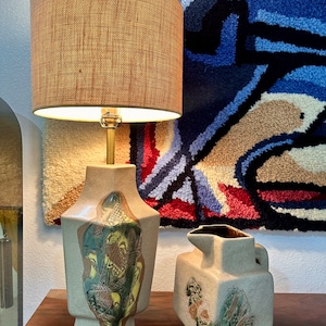 May include: A pair of ceramic lamps with a geometric design and a beige and brown color scheme. The lamps feature a stylized floral pattern with green, yellow, and brown accents. The lamps are on a wooden surface.