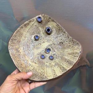 May include: A handmade, ceramic plate with a unique, organic shape. The plate is primarily tan with a dark brown rim and features several small, blue, circular indentations. The plate's surface has a textured, speckled appearance, suggesting a rustic aesthetic.