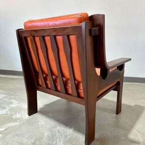 May include: A mid-century modern armchair with a dark wood frame and orange leather upholstery. The chair has a slatted back and a unique design with a curved armrest.