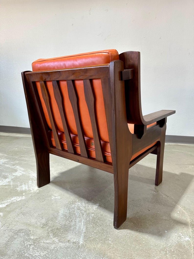 May include: A mid-century modern armchair with a dark wood frame and orange leather upholstery. The chair has a slatted back and a unique design with a curved armrest.