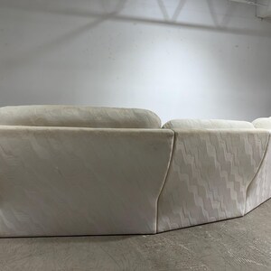 May include: Cream-colored sectional sofa with a geometric pattern. The sofa has a curved design and a low profile. The fabric has a subtle wave-like texture. The sofa is in a neutral color, suitable for various interior styles.