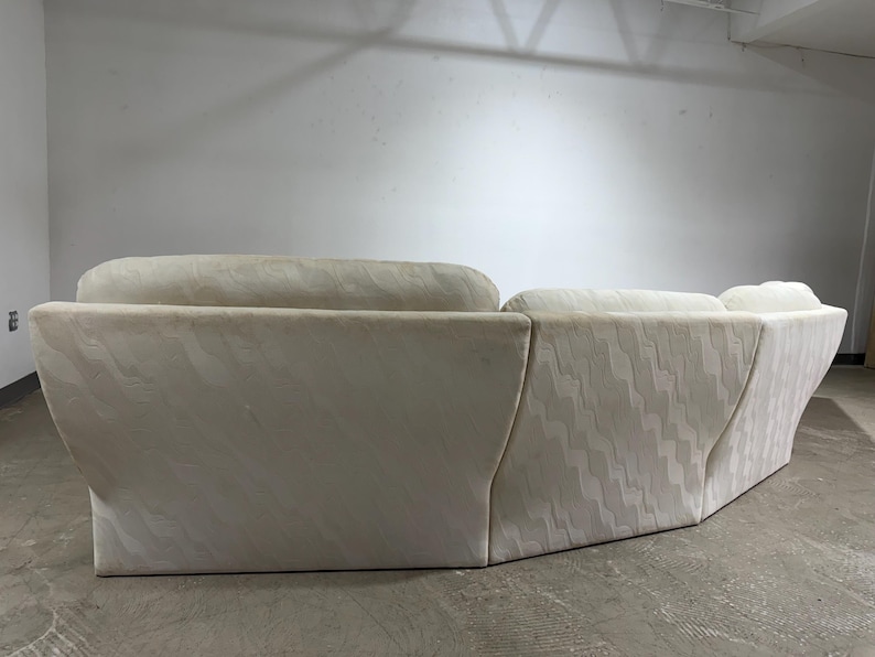 May include: Cream-colored sectional sofa with a geometric pattern. The sofa has a curved design and a low profile. The fabric has a subtle wave-like texture. The sofa is in a neutral color, suitable for various interior styles.
