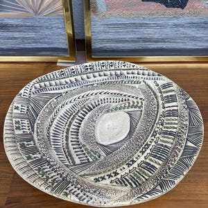 May include: A decorative ceramic plate with a spiral design. The plate features a cream-colored center and is encircled by intricate patterns in black, green, and cream. The plate is placed on a wooden surface, with artwork in the background.