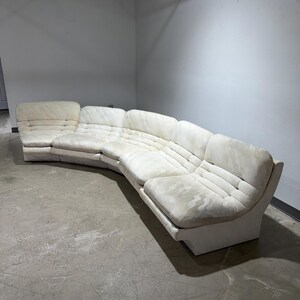 May include: A large, curved sectional sofa in a light cream color. The sofa has a low profile with a series of connected sections. The cushions have a quilted pattern. The sofa is in a room with a concrete floor and white walls.