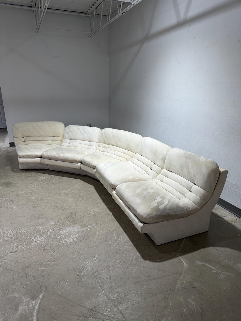 May include: A large, curved sectional sofa in a light cream color. The sofa has a low profile with a series of connected sections. The cushions have a quilted pattern. The sofa is in a room with a concrete floor and white walls.