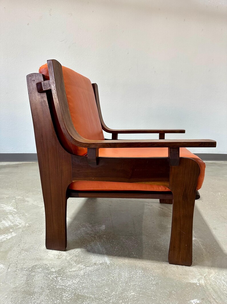 May include: A mid-century modern armchair with a dark wood frame and orange leather upholstery. The chair has a unique design with a high back and wide arms.