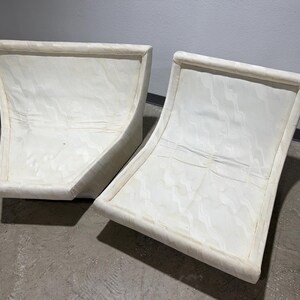 May include: Two off-white, curved-back chairs. The chairs have a rectangular frame with a curved back and seat. The fabric appears to have a subtle pattern. The chairs are placed on a concrete floor.