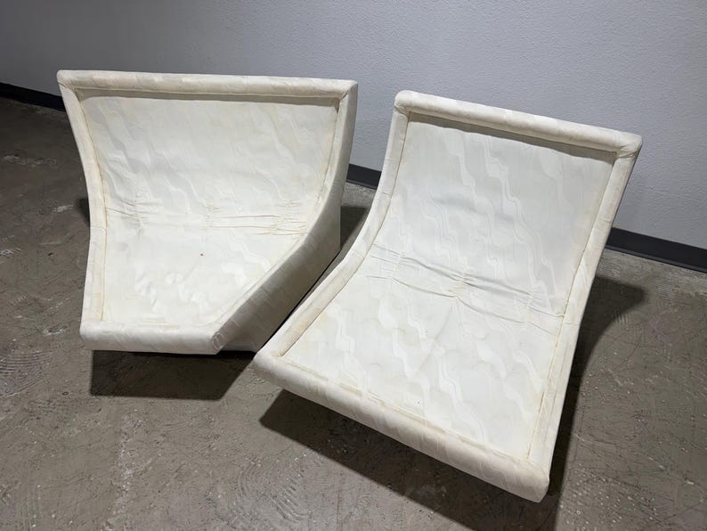 May include: Two off-white, curved-back chairs. The chairs have a rectangular frame with a curved back and seat. The fabric appears to have a subtle pattern. The chairs are placed on a concrete floor.