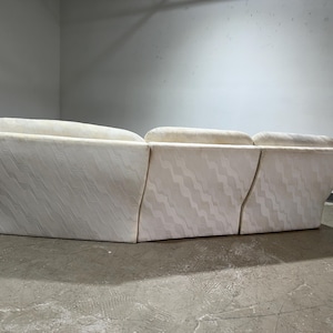 May include: A sectional sofa with a cream-colored fabric featuring a subtle, wavy pattern. The sofa has a low profile and a curved design, with multiple sections joined together. The background is a plain, light-colored wall.