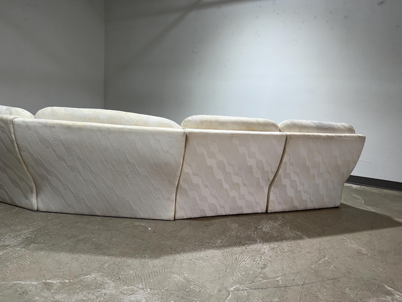 May include: A sectional sofa with a cream-colored fabric featuring a subtle, wavy pattern. The sofa has a low profile and a curved design, with multiple sections joined together. The background is a plain, light-colored wall.