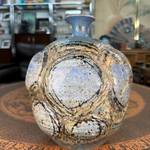 May include: A handcrafted ceramic vase with a bulbous body and a narrow neck. The vase features a textured surface with a mix of blue, tan, and black colors. The design includes circular patterns and a speckled finish, creating a unique, artistic look.