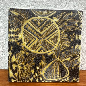 May include: A square ceramic tile with a black background and a raised yellow design. The design features a central circle with geometric patterns and surrounding floral motifs.