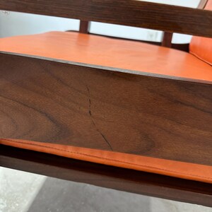 May include: Close-up of a wooden chair with an orange leather seat. The chair has a simple, mid-century modern design.