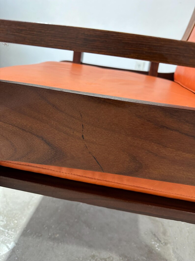 May include: Close-up of a wooden chair with an orange leather seat. The chair has a simple, mid-century modern design.