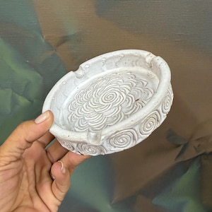 May include: A white ceramic ashtray with a floral design in the center and spiral patterns around the rim. The ashtray is round and has small grooves for resting cigarettes.
