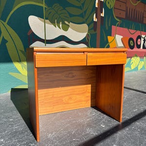 May include: A wooden desk featuring a warm, light brown grain finish. The desk has a rectangular top and two side-by-side drawers positioned directly beneath the surface. It is supported by two solid side panels that extend to the floor.