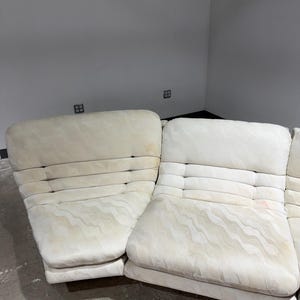 May include: Off-white sectional sofa with a patterned fabric. The sofa has a low profile with a series of rectangular cushions and a tufted back. The fabric has a subtle wave-like design. The sofa is in a room with gray walls.