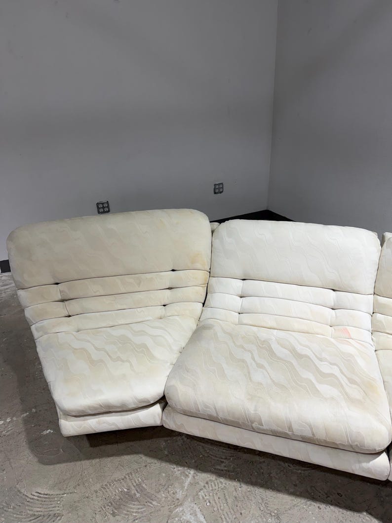 May include: Off-white sectional sofa with a patterned fabric. The sofa has a low profile with a series of rectangular cushions and a tufted back. The fabric has a subtle wave-like design. The sofa is in a room with gray walls.