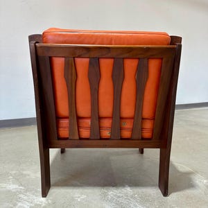 May include: A mid-century modern armchair with a dark wood frame and orange leather upholstery. The chair has a unique back design with vertical slats.