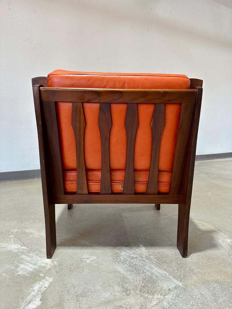 May include: A mid-century modern armchair with a dark wood frame and orange leather upholstery. The chair has a unique back design with vertical slats.