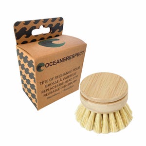 May include: A round bamboo dish brush head with light-coloured bristles next to a brown cardboard box. The box features a black wave pattern, the text OCEANSRESPECT, and the words TÊTE DE RECHANGE POUR BROSSE À VAISSELLE REUSABLE DISH BRUSH.