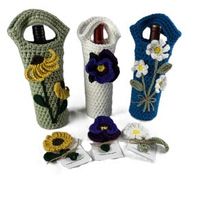 May include: Three handmade crochet wine bottle covers in green, white, and blue, each adorned with a different flower: sunflower, pansy, and daisy. Each cover has a handle. Additional flower pins are included.