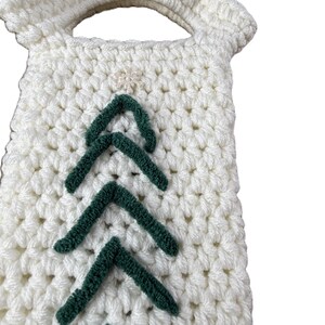 Pu&ograve; includere: Una borsa all'uncinetto color crema con manico e un motivo verticale di galloni in lana verde scuro disposti a forma di albero. Un piccolo decoro bianco a forma di stella &egrave; centrato nella parte superiore del motivo a galloni.