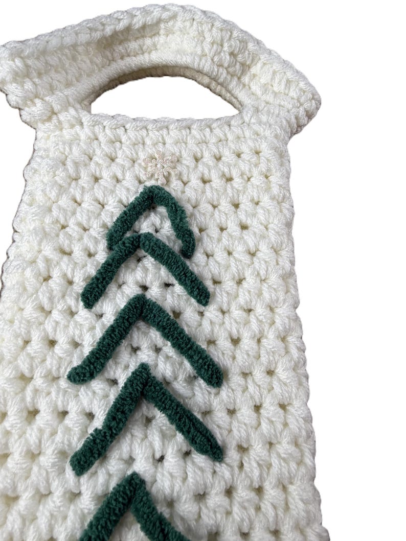 Pu&ograve; includere: Una borsa all'uncinetto color crema con manico e un motivo verticale di galloni in lana verde scuro disposti a forma di albero. Un piccolo decoro bianco a forma di stella &egrave; centrato nella parte superiore del motivo a galloni.