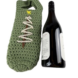 Pu&ograve; includere: Una custodia per bottiglia di vino all'uncinetto color verde salvia con un tronco d'albero in filo marrone, rami in filo bianco e una piccola stella in filo giallo in cima. Accanto alla custodia c'&egrave; una bottiglia di vino in vetro scuro.