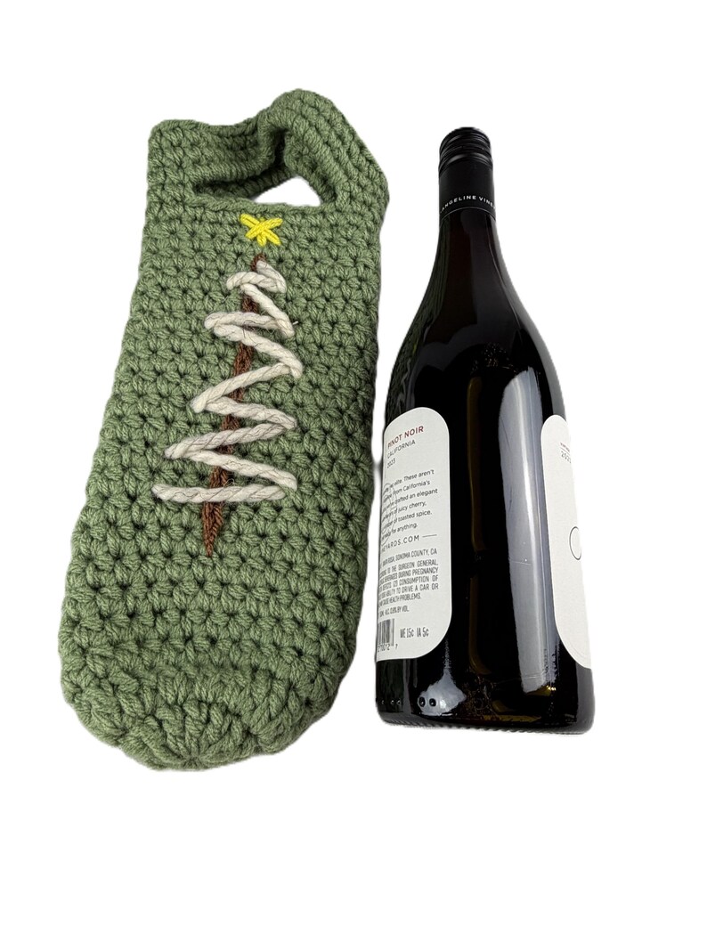 Pu&ograve; includere: Una custodia per bottiglia di vino all'uncinetto color verde salvia con un tronco d'albero in filo marrone, rami in filo bianco e una piccola stella in filo giallo in cima. Accanto alla custodia c'&egrave; una bottiglia di vino in vetro scuro.