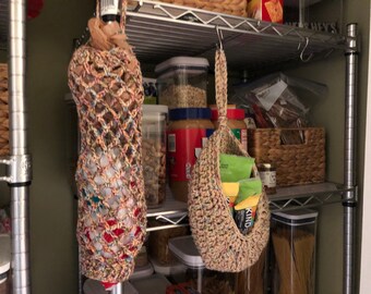 May include: Two crocheted hanging bags in a pantry. The bag on the left is filled with a clear plastic bottle. The bag on the right is filled with snack bars. Both bags are made with multi-colored yarn.